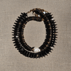 Close-up of LUMÉA bold coconut disc necklace featuring layered black and dark brown coconut beads, gold-tone accents, and a central white baroque pearl on a textured beige background