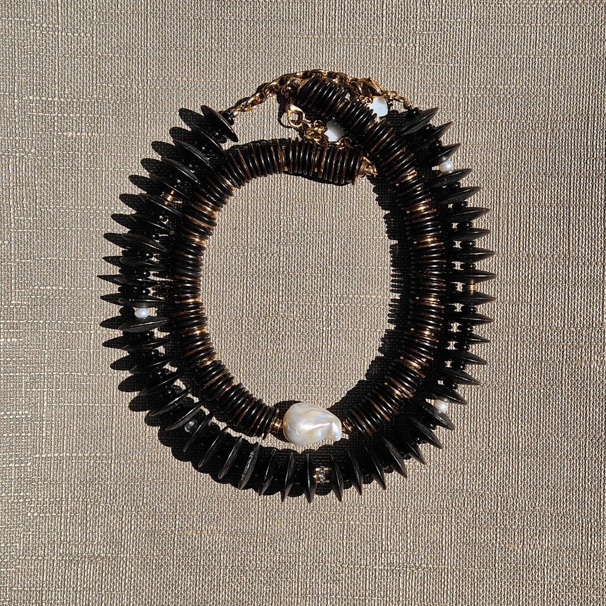 Close-up of LUMÉA bold coconut disc necklace featuring layered black and dark brown coconut beads, gold-tone accents, and a central white baroque pearl on a textured beige background