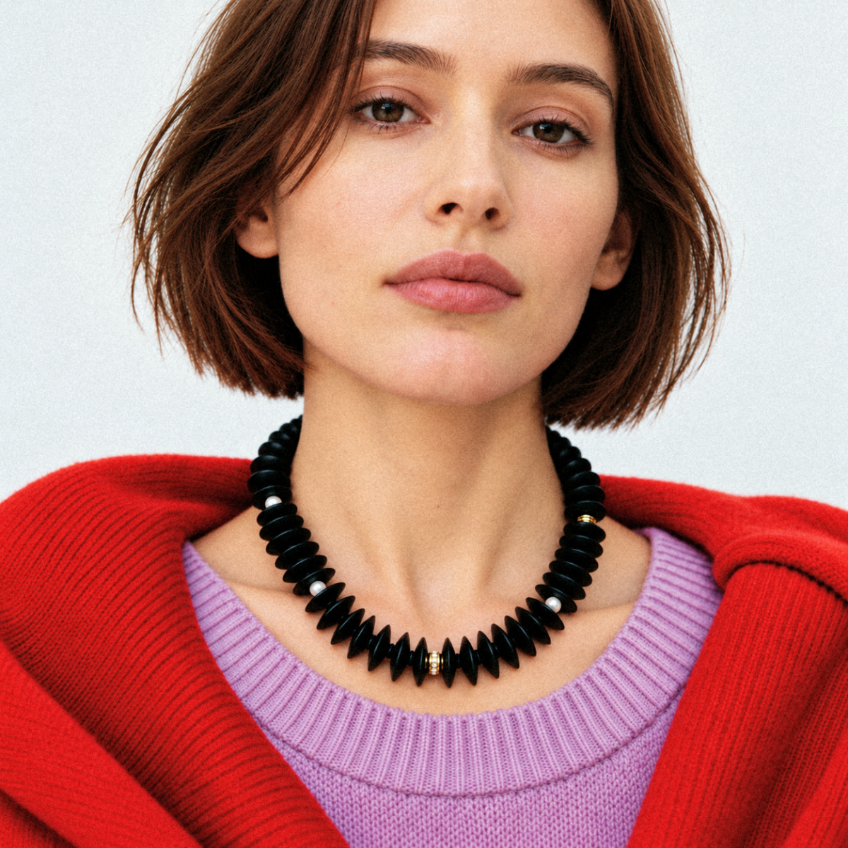 Model wearing LUMÉA bold black coconut disc necklace with gold-tone accents, styled with a lavender knit sweater and red outerwear for a modern fall and winter look.