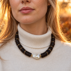 Model wearing a bold LUMÉA coconut disc and baroque pearl necklace with dark brown and black beads, styled over a cream knit sweater for fall and winter.